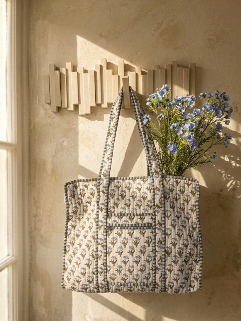 A quilted tote bag with a beige and gray floral pattern hangs on a wooden wall rack against a textured beige wall, with blue flowers tucked into the side.