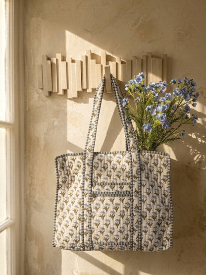 A quilted tote bag with a beige and gray floral pattern hangs on a wooden wall rack against a textured beige wall, with blue flowers tucked into the side.